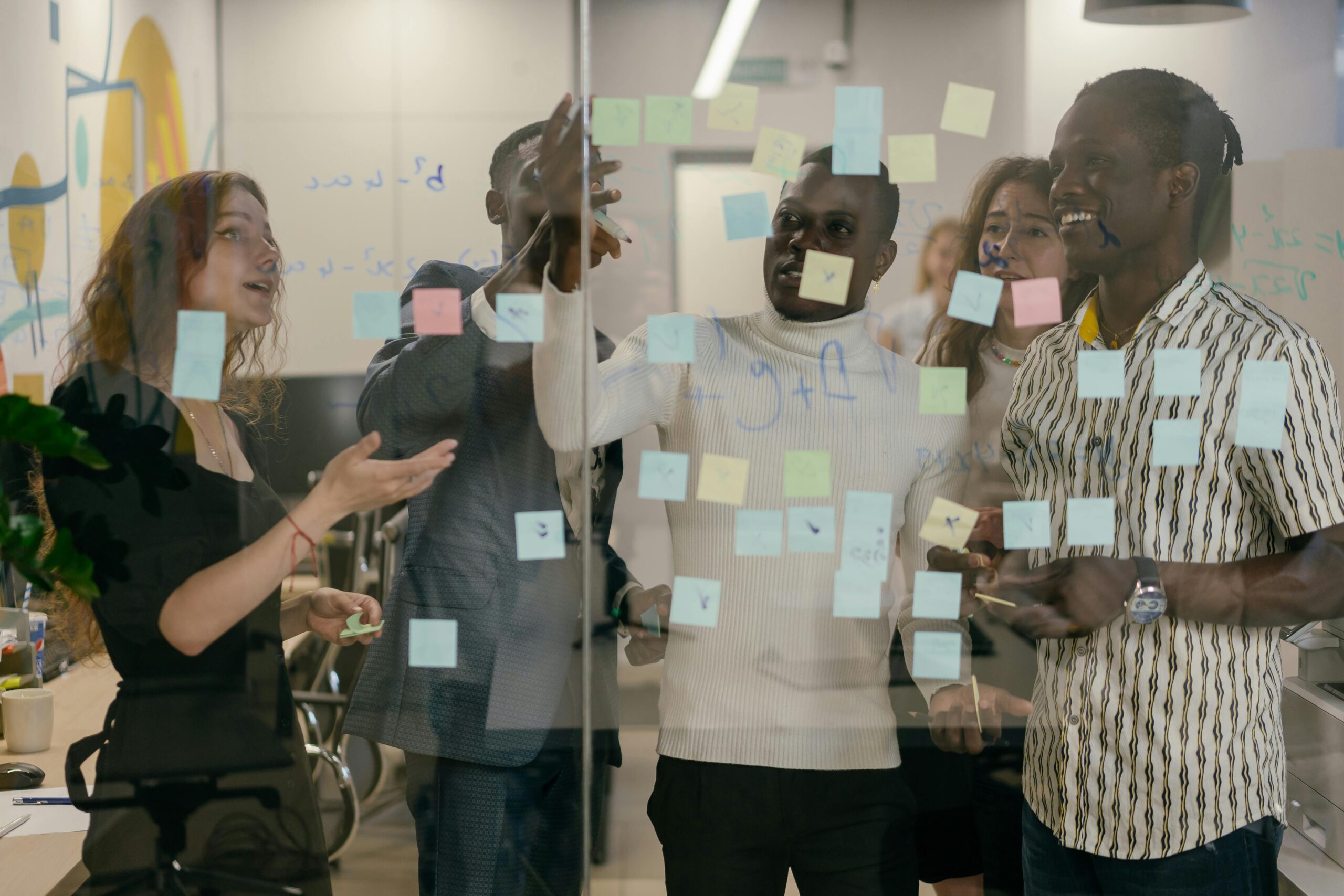 A group of five people gathered in an office in front of a glass panel with sticky notes discussing a topic as an employee resource group (ERG)