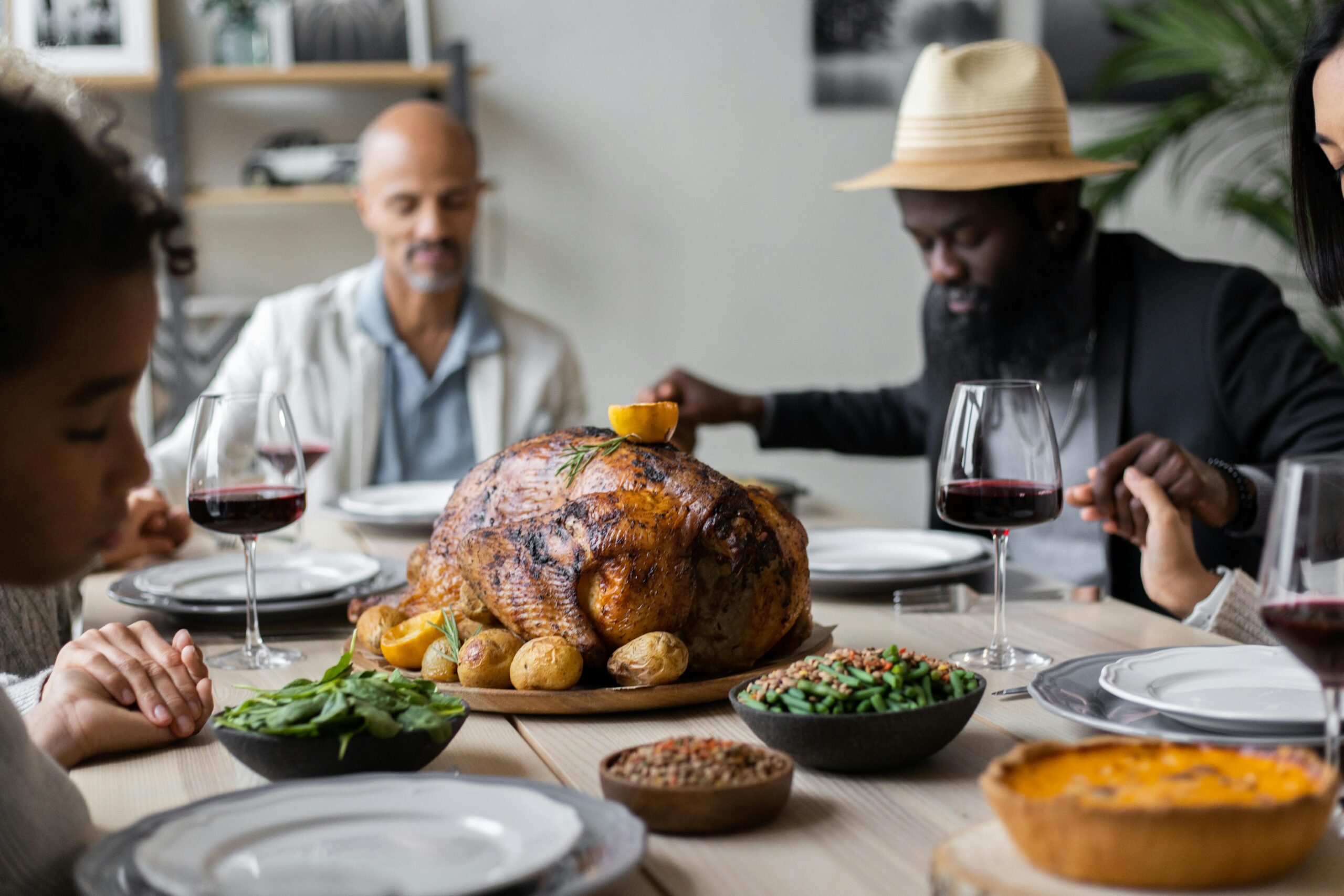 A family holding hands with their heads bowed and eyes closed at the dinner table over a thanksgiving turkey.